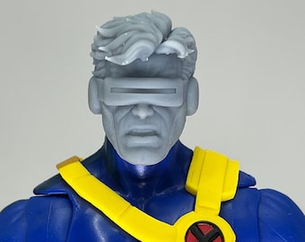 May include: A blue and yellow action figure with a gray head and visor. The figure has a red X on its chest.