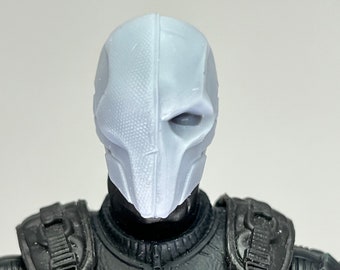 May include: A white plastic action figure head with a detailed mask. The mask has a single eye hole and a textured surface.