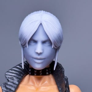 Könnte beinhalten: Detaillierte Nahaufnahme eines Puppenkopfes und des Oberkörpers. Die Puppe hat hellblaue Haut, weißes Haar und einen schwarzen, mit Nieten besetzten Halsband. Die Puppe trägt eine schwarze Jacke und ein orangefarbenes Oberteil.