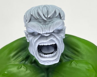 May include: A close-up of a grey, sculpted head of the Hulk with a wide-open mouth and a fierce expression. The head is set against a green background.