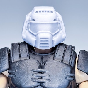 Puede incluir: Una figura de acción gris claro con cabeza y armadura de pecho. El casco tiene una visera y ranuras de ventilación. La armadura del pecho es negra con paneles detallados. La piel de la figura es de color canela claro.