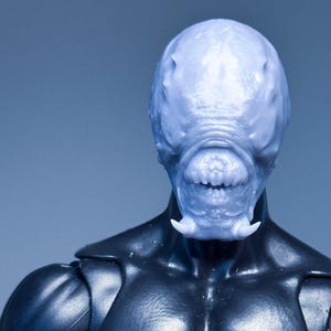May include: A light blue, textured creature head with a smooth, rounded cranium, a wide mouth filled with small teeth, and two prominent, downward-pointing fangs, attached to a black, muscular torso with defined chest and shoulder musculature.