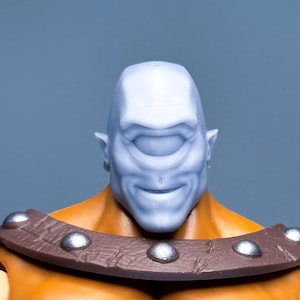 以下が含まれることがあります： ライトブルーの頭と単眼を持つサイクロプスのアクションフィギュアのクローズアップ。フィギュアは、シルバーのスタッドが付いた茶色の襟とオレンジ色の胴体を持っています。背景は無地の青です。