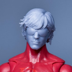 Puede incluir: Una cabeza de figura de acción de resina gris claro sin pintar con un peinado texturizado y gafas de sol moldeadas. La cabeza está unida a un torso rojo brillante con una letra D tenue visible en el pecho, mostrando rasgos faciales detallados.