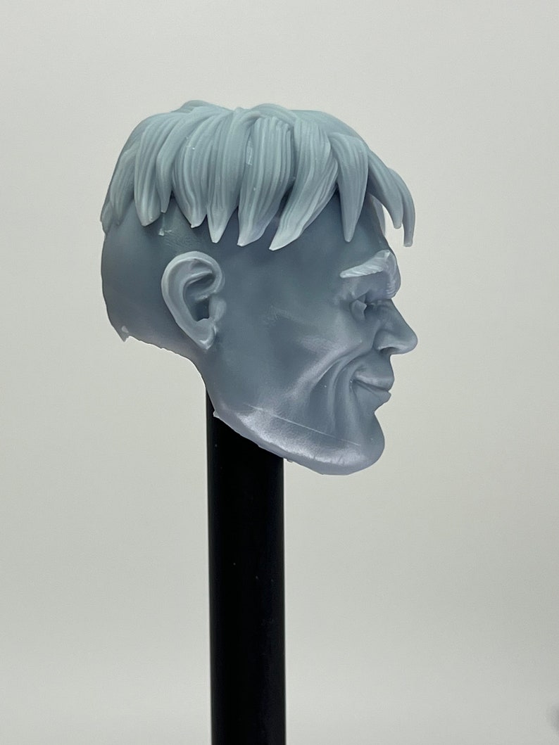 May include: A gray 3D printed head sculpt of a man with short, textured hair and a slight smile. The head is mounted on a black stand.