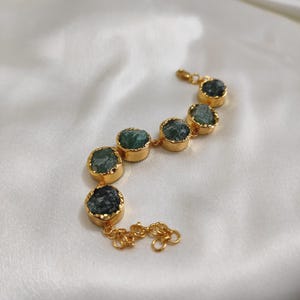 May include: Gold-toned bracelet featuring seven rough-cut, emerald-green gemstones. Each stone is set in a gold bezel and linked by delicate gold chain. The bracelet is displayed on a white, silky surface.