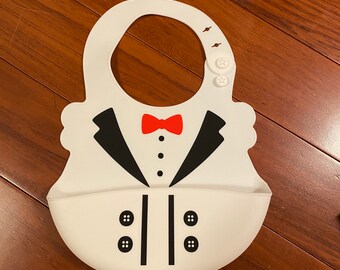 Puede incluir: Un babero de silicona blanco con un diseño de esmoquin impreso que incluye una pajarita roja, solapas negras, tres botones negros en el pecho y cuatro botones negros en la parte inferior. El babero cuenta con un bolsillo recogemigas y cierres de presión en forma de estrella.