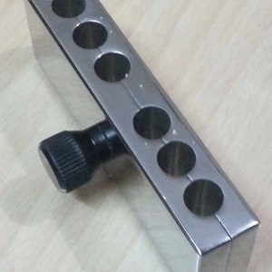 Peut inclure: Un outil en métal argenté avec six trous ronds et un bouton noir. L'outil est utilisé pour maintenir et aligner des objets.