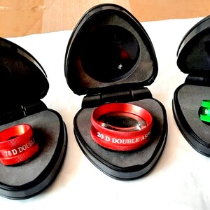 Op de afbeelding: Drie rode en groene ophthalmische lenzen in individuele zwarte koffers. De lenzen zijn gelabeld "7,8 D Double Aspheric", "20 D Double Aspheric" en "9,0 Double Aspheric".
