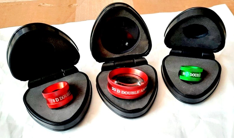 Op de afbeelding: Drie rode en groene ophthalmische lenzen in individuele zwarte koffers. De lenzen zijn gelabeld "7,8 D Double Aspheric", "20 D Double Aspheric" en "9,0 Double Aspheric".