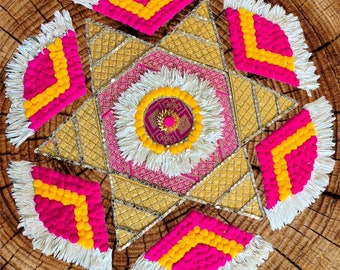 May include: A decorative, star-shaped textile art piece with a central floral design. The piece features gold, pink, and white elements, with accents of yellow and fringe. The background is a textured, wooden surface.