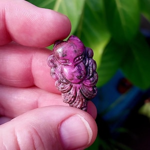 May include: A carved pink gemstone pendant in the shape of a mythical creature with intricate details. The pendant is made of a polished stone with a smooth, glossy finish.