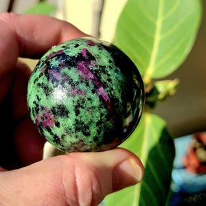 May include: A polished gemstone sphere, primarily green with black and magenta inclusions. The sphere is held in a hand, with a bright green leaf in the background. The gemstone has a smooth, reflective surface.