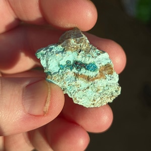 May include: A close-up of a mineral specimen held between fingers. The stone features a vibrant turquoise hue with patches of brown and dark blue. The texture appears rough and natural, showcasing the raw beauty of the mineral.