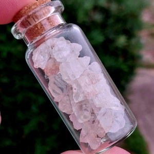 May include: A small glass bottle with a cork stopper filled with small, clear, and translucent crystals. The crystals have a slight pink hue.