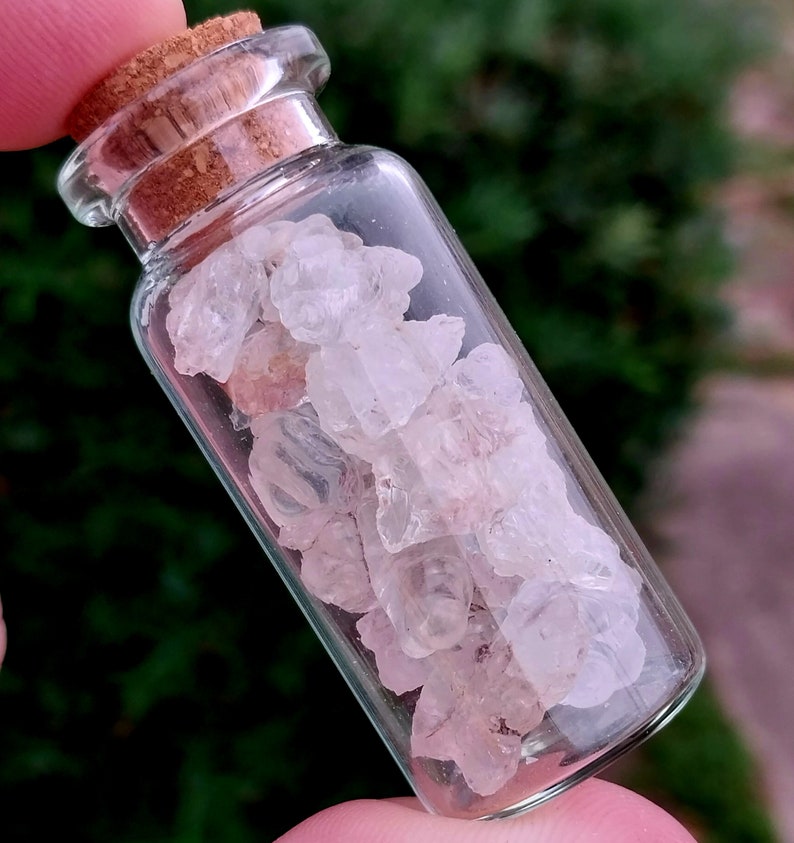 May include: A small glass bottle with a cork stopper filled with small, clear, and translucent crystals. The crystals have a slight pink hue.