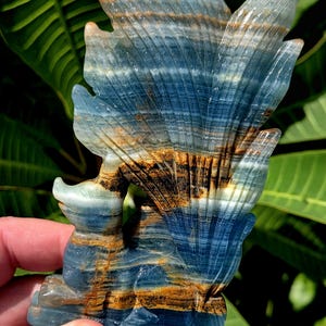 May include: A carved stone sculpture of a bird-like creature with blue, brown, and white layered patterns. The sculpture has a fan-shaped tail and is held in a person's hand. The background is blurred green foliage.