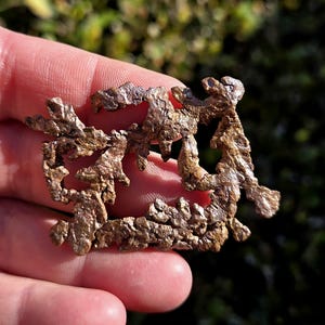 May include: A close-up of a raw, irregularly shaped copper nugget. The nugget has a rough, textured surface with a mix of brown and reddish-brown hues. The natural formation is held in a person's hand, with a blurred green background.