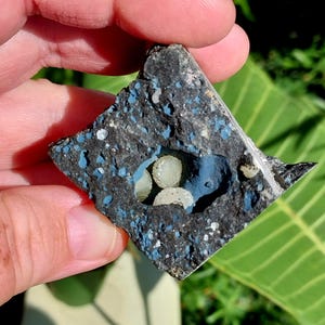May include: A piece of dark gray rock with blue speckles and a central cavity. The cavity is painted blue and contains several small, light-colored, spherical objects. The rock is held in a person's hand.