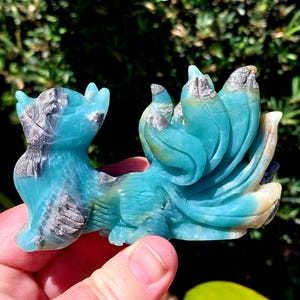 May include: A blue and white carved stone figurine of a mythical creature with a long, flowing tail. The creature has a small head with a pointed snout and large, round eyes. The tail is curled up behind the creature's body.