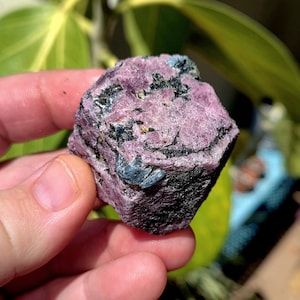 May include: A rough, uncut ruby gemstone with a deep pink hue, accented by black and dark blue mineral inclusions. The natural crystal formation is held in a hand, showcasing its raw, textured surface and unique color variations.