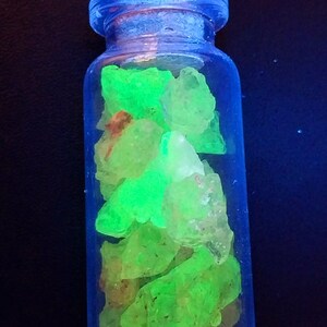 May include: A small glass bottle filled with bright green, fluorescent crystals. The bottle has a clear glass body and a blue lid.