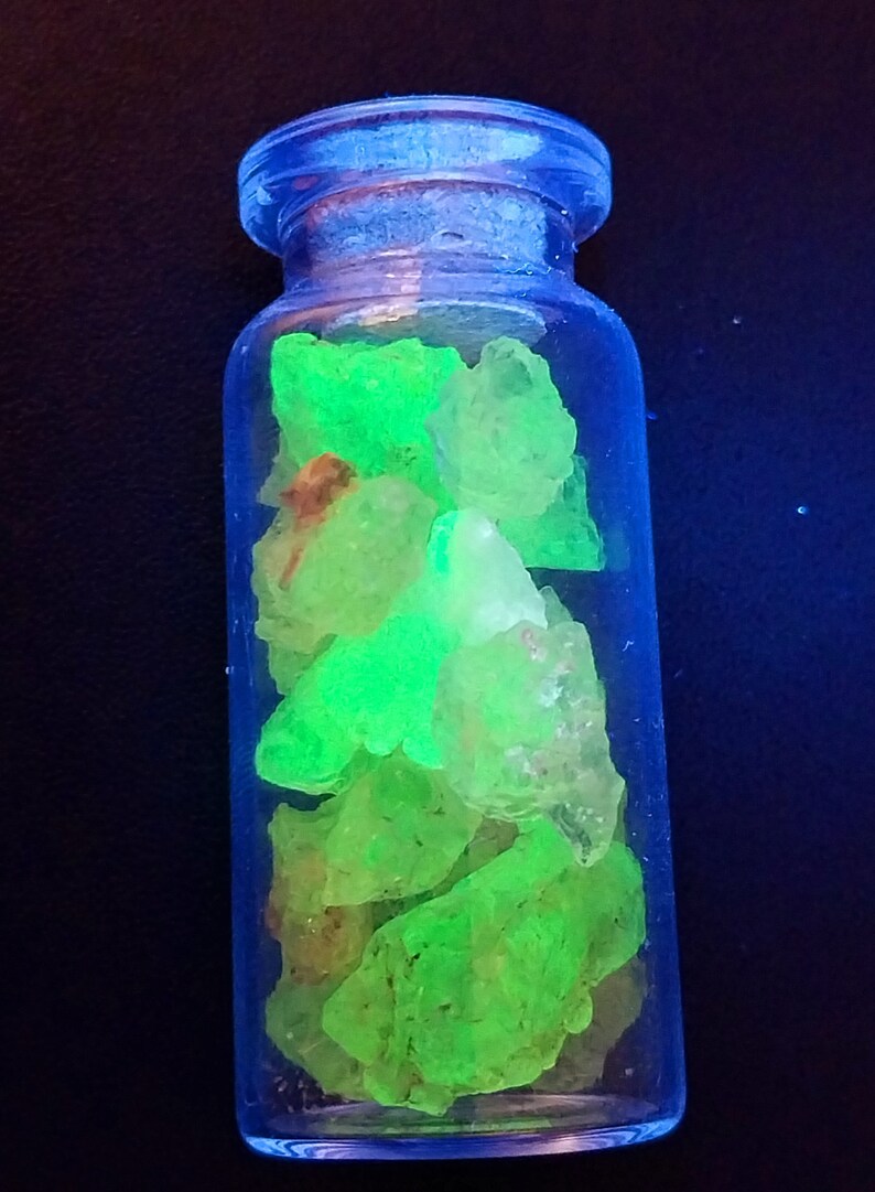 May include: A small glass bottle filled with bright green, fluorescent crystals. The bottle has a clear glass body and a blue lid.