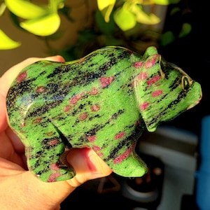 May include: A carved bear figurine made of green zoisite with black and pink inclusions. The bear is in a standing position, with its features carefully sculpted. The gemstone has a polished finish, enhancing the colors and details of the bear.