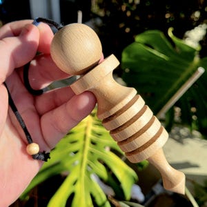 May include: A light-colored wooden pendulum with a round top and a carved, ridged body. A black cord with a wooden bead is attached to the top. The pendulum is held against a blurred background of green leaves.