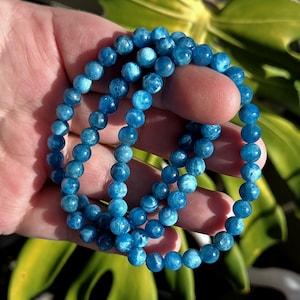 May include: A strand of blue apatite gemstone beads. The beads are a vibrant shade of blue with a slightly marbled appearance. The bracelet is held in a hand, with a blurred green background.