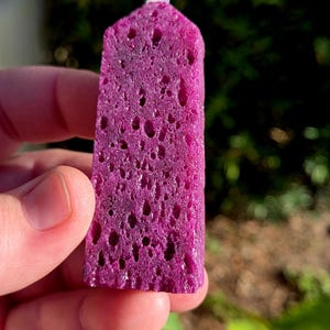 May include: A vibrant magenta crystal point with a porous texture, held in a hand. The crystal has a rectangular shape with a rounded top and a small, white, spherical accent. The background is blurred green foliage.