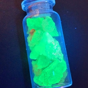 May include: A glass bottle filled with green, glowing crystals. The bottle has a cork stopper and is made of clear glass.