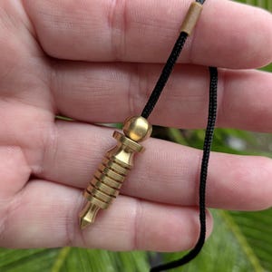 May include: A brass pendulum with a black cord. The pendulum has a cylindrical body with horizontal grooves and a pointed tip. The cord is attached to a small, round brass bead at the top.