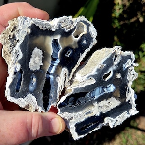 May include: Two pieces of a polished stone with a striking pattern. The stone features layers of black, white, and gray, with a rough, textured exterior. The interior reveals intricate banding and natural formations, creating a unique and artistic appearance.