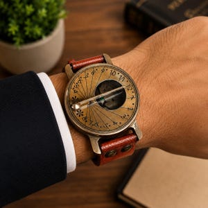 May include: A brass sundial watch with a circular face featuring engraved numbers and a central gnomon, attached to a textured brown leather strap with two metal rivets. The watch dial displays a radial pattern with markings for timekeeping.