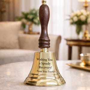 Pode incluir: Um sino de latão com uma alça de madeira marrom escura e um botão dourado. O sino tem a inscrição "Wishing You a Speedy Recovery! - From Your Friends" em texto preto. O sino está em uma mesa de mármore.