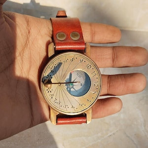 May include: A vintage brass sundial watch with a red leather strap. The watch face features a compass and hour markings. The watch is held in a hand, showcasing its size and design. A unique and functional accessory.
