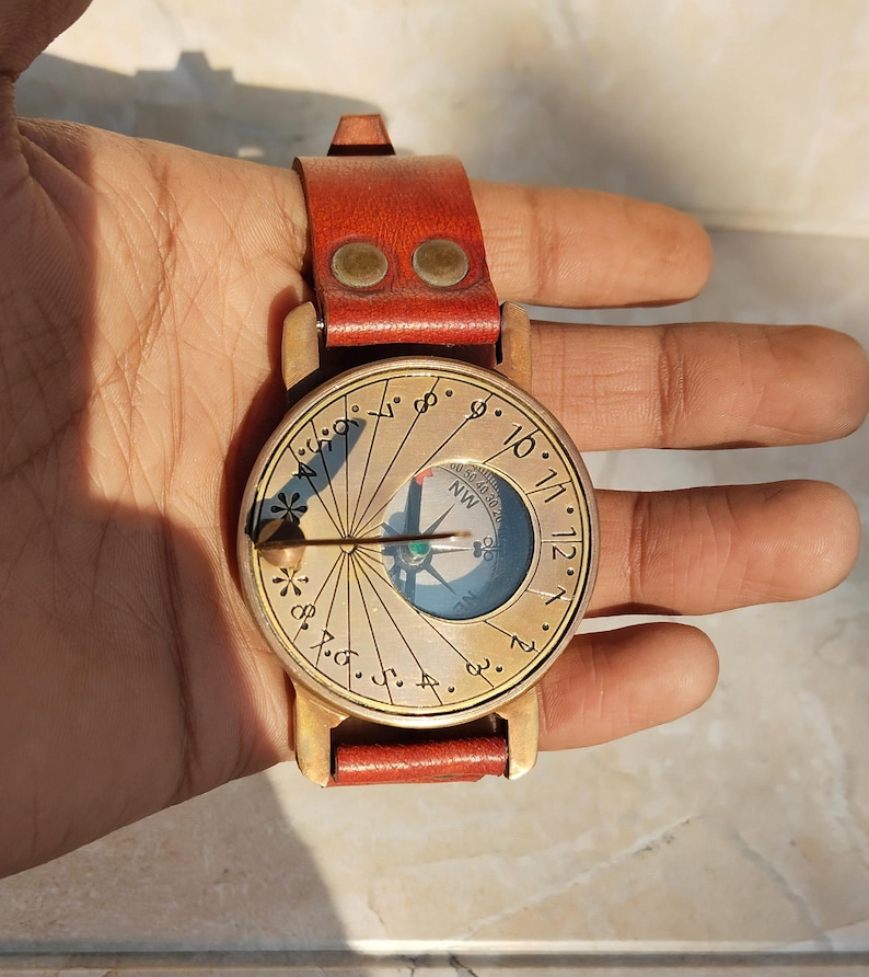 May include: A vintage brass sundial watch with a red leather strap. The watch face features a compass and hour markings. The watch is held in a hand, showcasing its size and design. A unique and functional accessory.