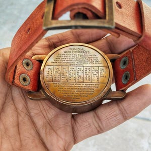 May include: A vintage sun dial and compass with a leather strap. The brass dial features a time conversion table and the text "SUN DIAL AND COMPASS." The leather strap is reddish-brown with brass hardware.
