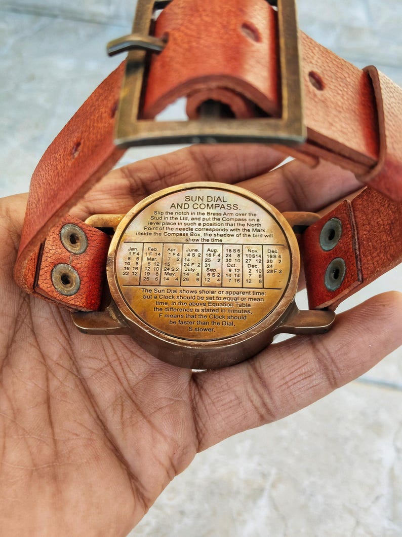 May include: A vintage sun dial and compass with a leather strap. The brass dial features a time conversion table and the text "SUN DIAL AND COMPASS." The leather strap is reddish-brown with brass hardware.