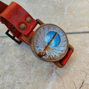 May include: A vintage-style compass watch with a red leather strap. The watch face has a blue center and a silver-colored outer ring with numbers and lines. The strap is secured with two rivets and a buckle. The watch is designed to be worn on the wrist.