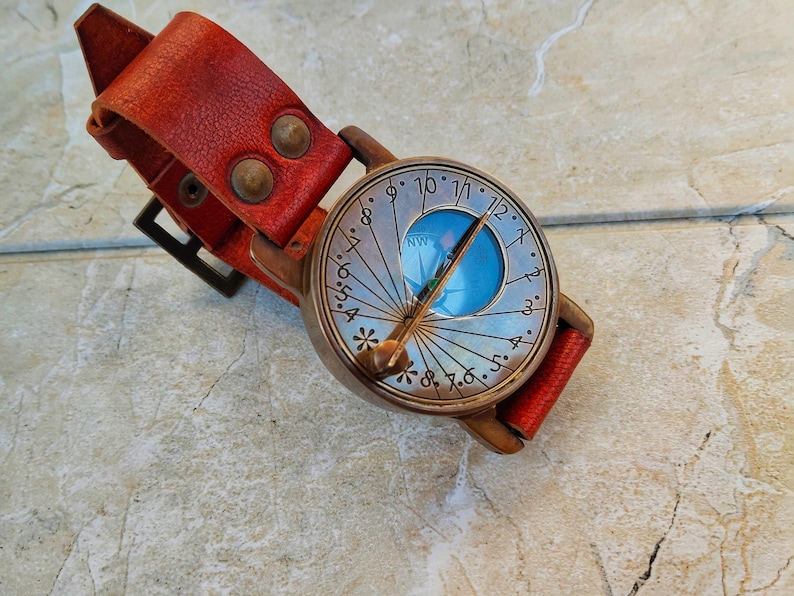 May include: A vintage-style compass watch with a red leather strap. The watch face has a blue center and a silver-colored outer ring with numbers and lines. The strap is secured with two rivets and a buckle. The watch is designed to be worn on the wrist.