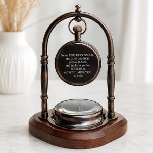 Puede incluir: Un reloj de escritorio de metal color bronce con una esfera circular suspendida de un marco arqueado sobre una base de madera oscura. La esfera muestra el texto: Never UNDERESTIMATE the DIFFERENCE you've MADE and the lives you've TOUCHED. WE WILL MISS YOU, ANNA.