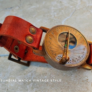 May include: Vintage-style sundial watch with a red leather strap. The watch face is bronze-colored with etched numerals and a gnomon. The strap has brass rivets and a buckle. The text "SUNDIAL WATCH VINTAGE STYLE" is at the bottom.