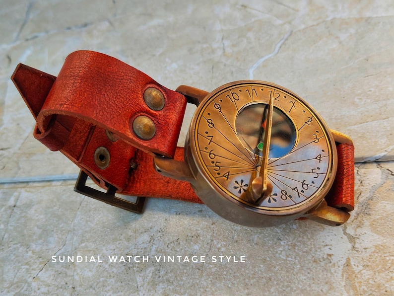 May include: Vintage-style sundial watch with a red leather strap. The watch face is bronze-colored with etched numerals and a gnomon. The strap has brass rivets and a buckle. The text "SUNDIAL WATCH VINTAGE STYLE" is at the bottom.