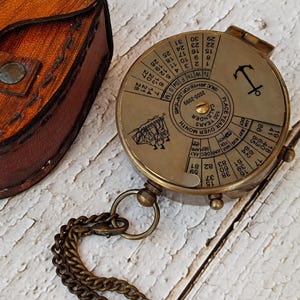 May include: A brass compass with a chain and a leather pouch. The compass has a calendar dial with the months of the year and days of the week. The compass also has a nautical anchor symbol.