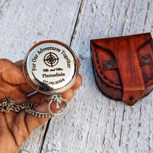 May include: A silver compass with a chain, engraved with "For Our Adventures Together," a compass rose, and the names "Mr. and Mrs. Plamadiala" with the date "07.09.2024." A brown leather case is next to the compass.