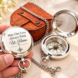 Peut inclure: Une boussole de poche argentée avec un couvercle à charnière gravé du texte 'May Our Love Always Point Us Home' et une rose des vents. Une seconde boussole ouverte révèle un cadran détaillé avec les points cardinaux et une chaîne en métal.