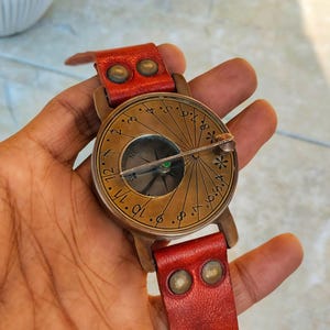 May include: A vintage-style compass watch with a brass face and a red leather strap. The compass features a detailed dial with directional markings and a needle. The watch has a classic, antique aesthetic.