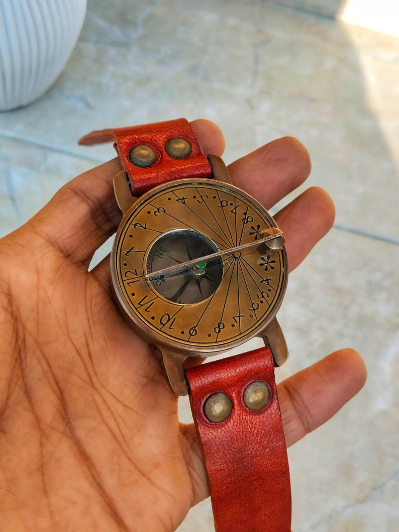 May include: A vintage-style compass watch with a brass face and a red leather strap. The compass features a detailed dial with directional markings and a needle. The watch has a classic, antique aesthetic.